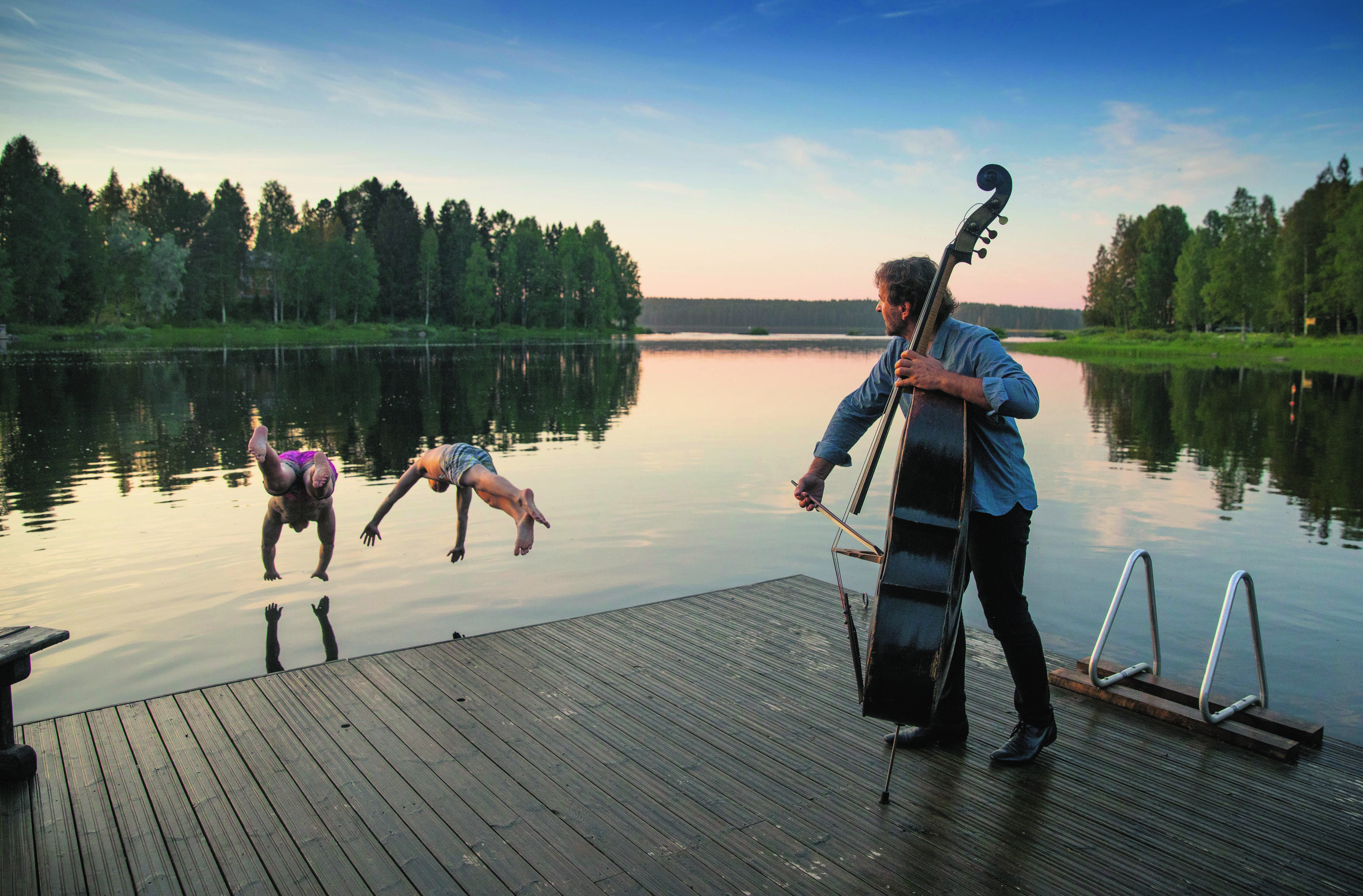 Sur un ponton, un homme joue du violoncelle en regardant des gens plonger dans l’eau dans la région des lacs